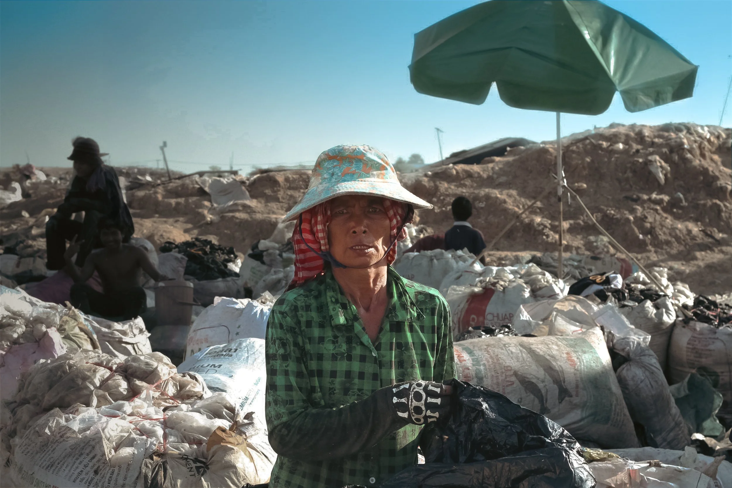 Une femme avec un chapeau à larges bords, portant une chemise à carreaux verte, se tient devant une grande quantité de sacs remplis de déchets ou de matériaux recyclés dans un site en plein air, sous un parasol vert, avec d'autres personnes en arrière-plan.