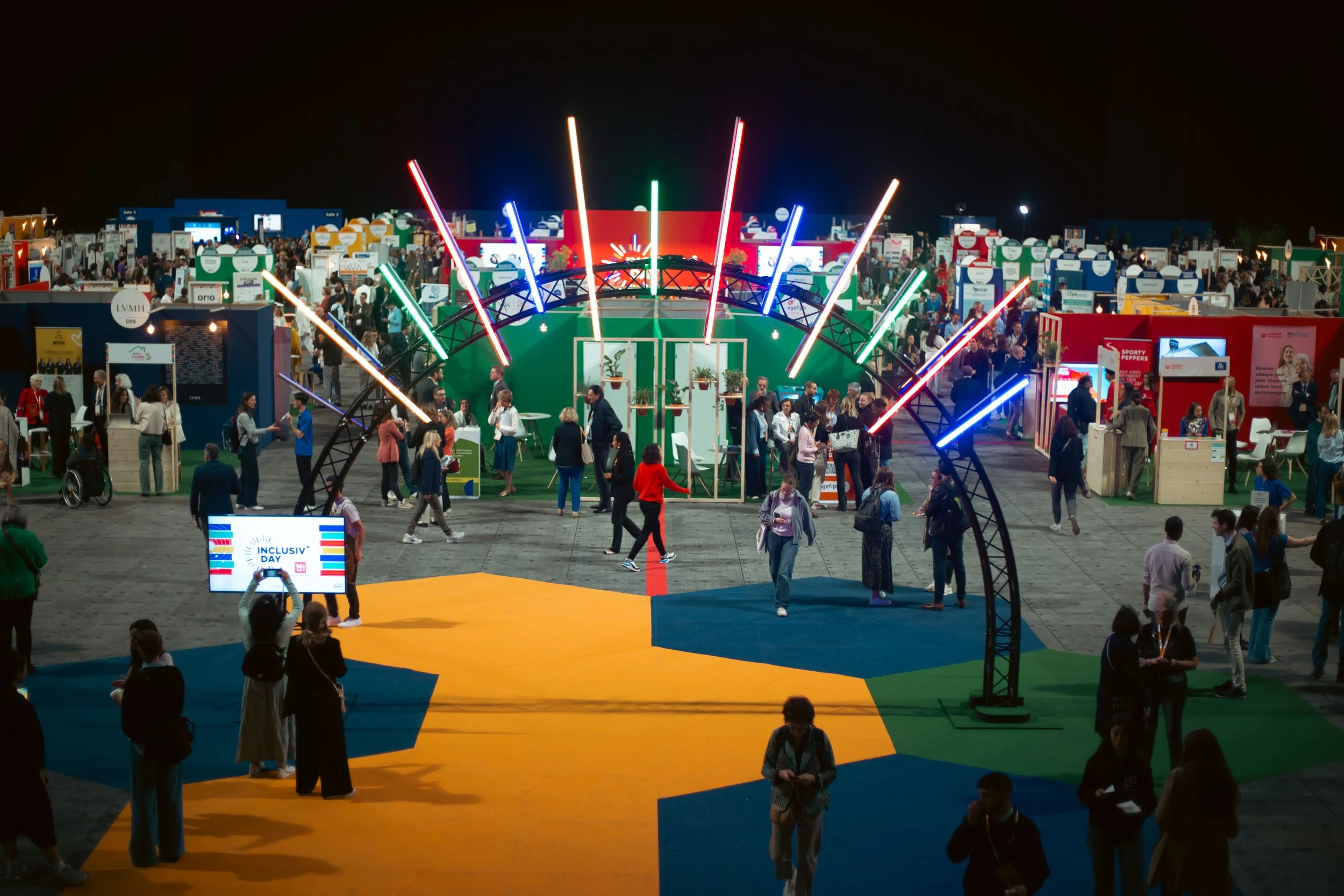 Vue d'ensemble d'un salon ou d'une exposition avec de nombreux stands. Au centre, un grand cerceau décoré de lumières colorées avec des personnes qui se déplacent et interagissent. Le sol est peint en plusieurs couleurs avec un panneau indiquant 'Inc