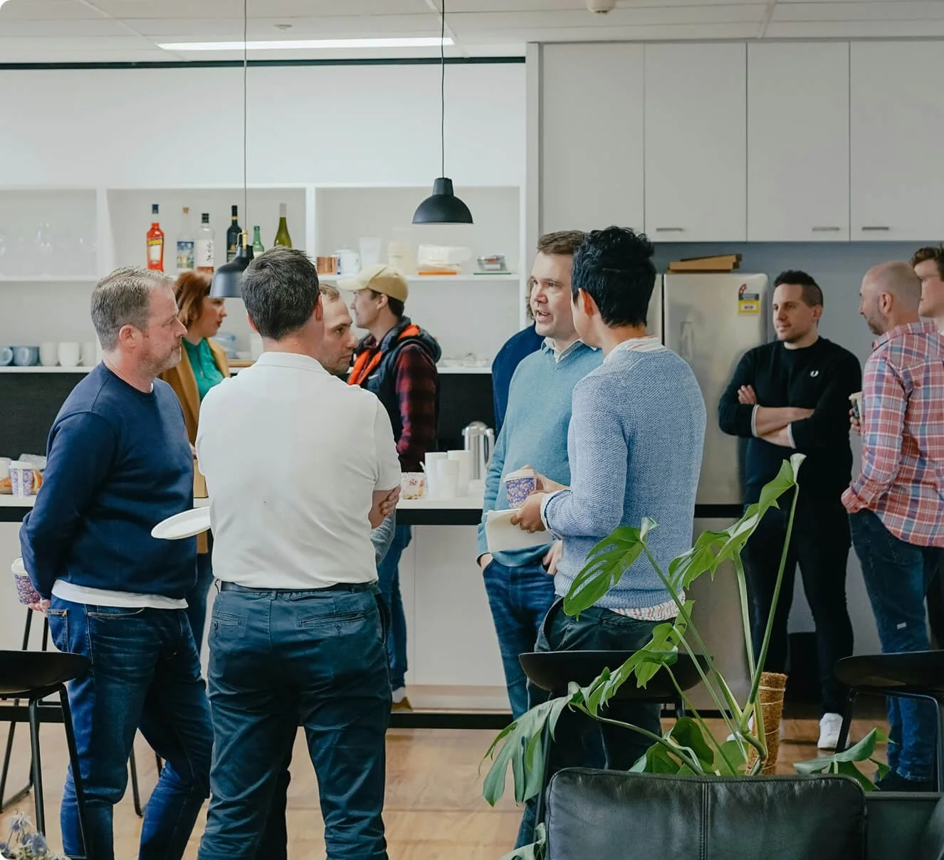 People socializing and chatting in a modern kitchen or break room, with drinks and snacks on the counter, and a plant in the foreground.