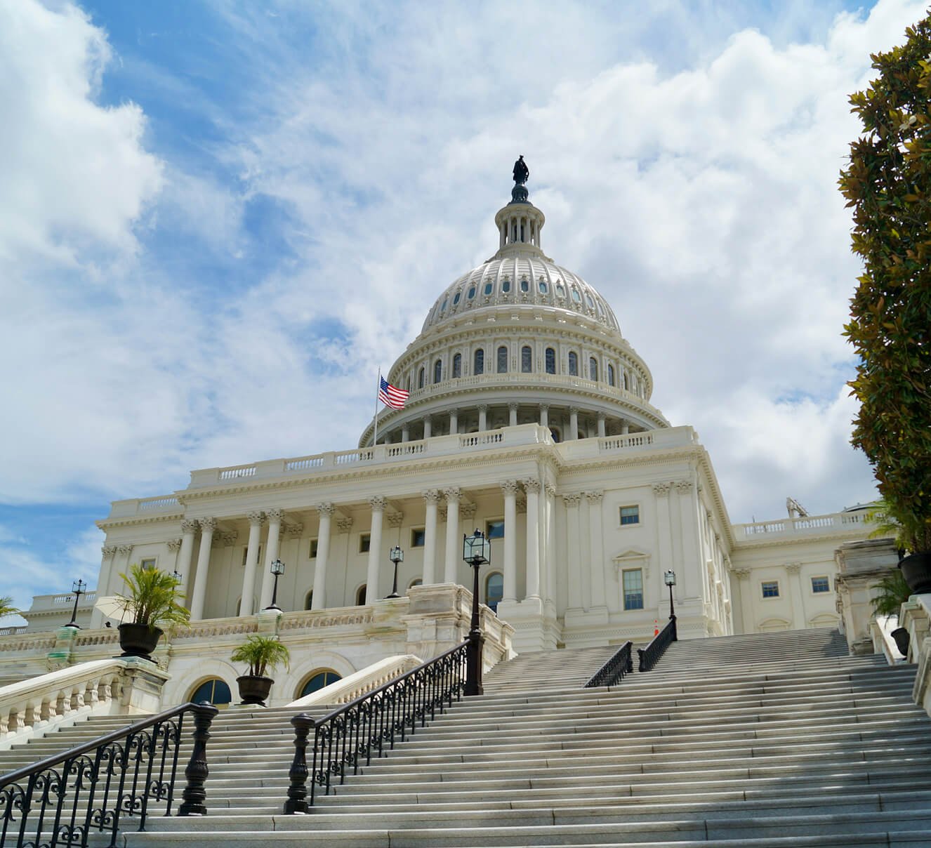 The United States Capitol building in Washington, D.C., with a prominent white dome, steps leading up to the entrance, and a flag flying near the top, under a partly cloudy sky.