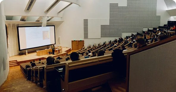 A lecture hall filled with students seated in tiered rows, facing a large projection screen and lectern at the front.