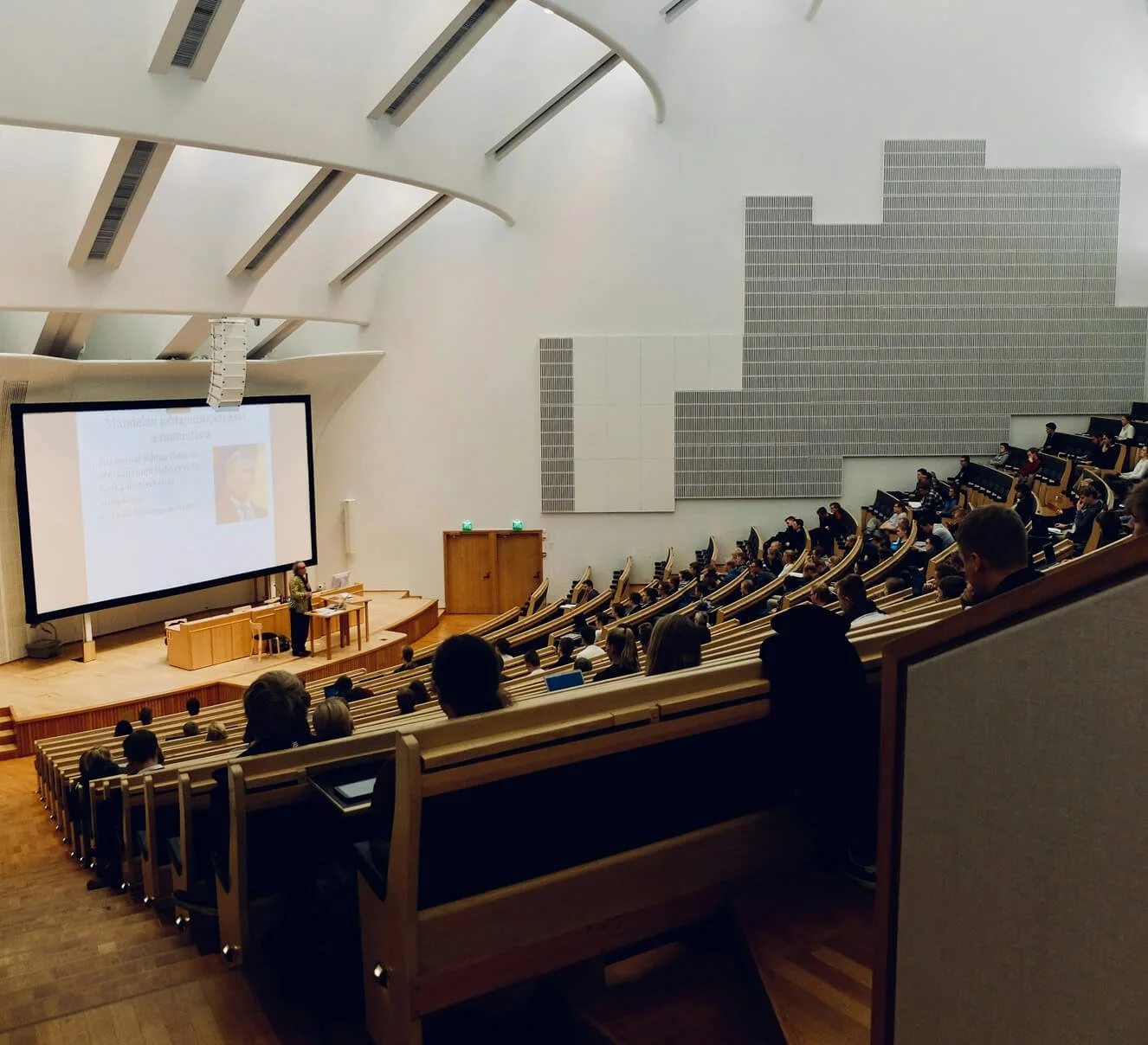 Lecture hall filled with students, a presenter standing at the podium, large screen displaying a slide, modern design, wood seating, some students using laptops.