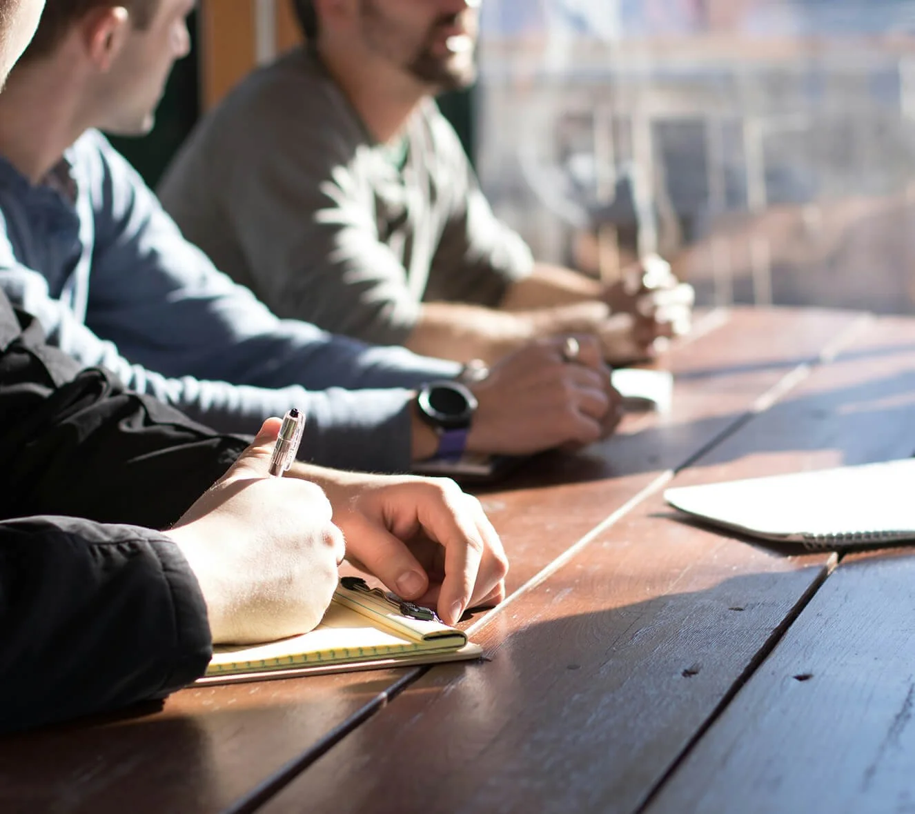 Three people sitting at a wooden table, with one person taking notes in a notepad, while the other two are seated beside them, near a large window letting in natural light.