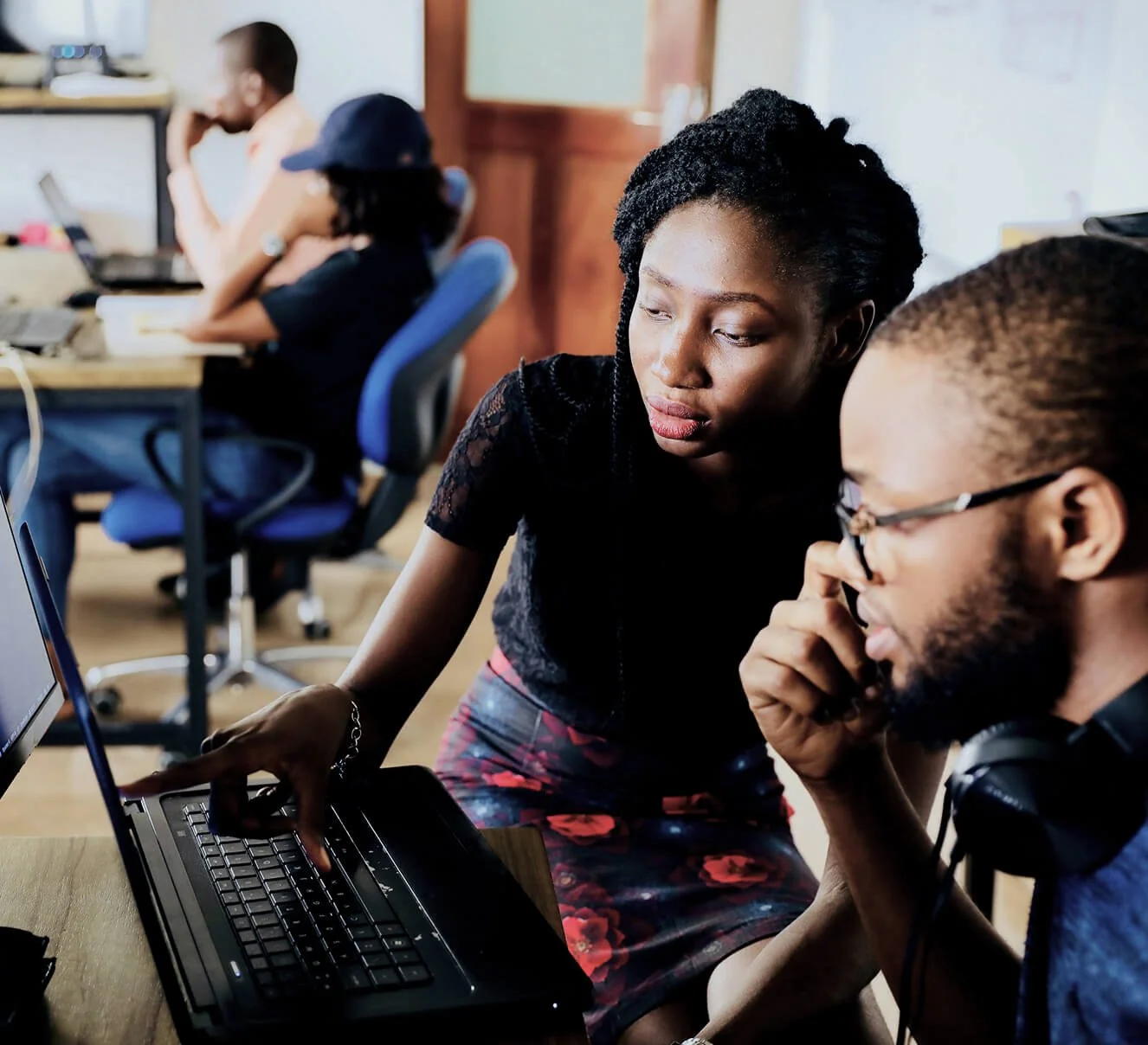 Two women are working on a laptop, with one woman pointing at the screen while the other woman is looking at it thoughtfully. In the background, there are other people working at desks.