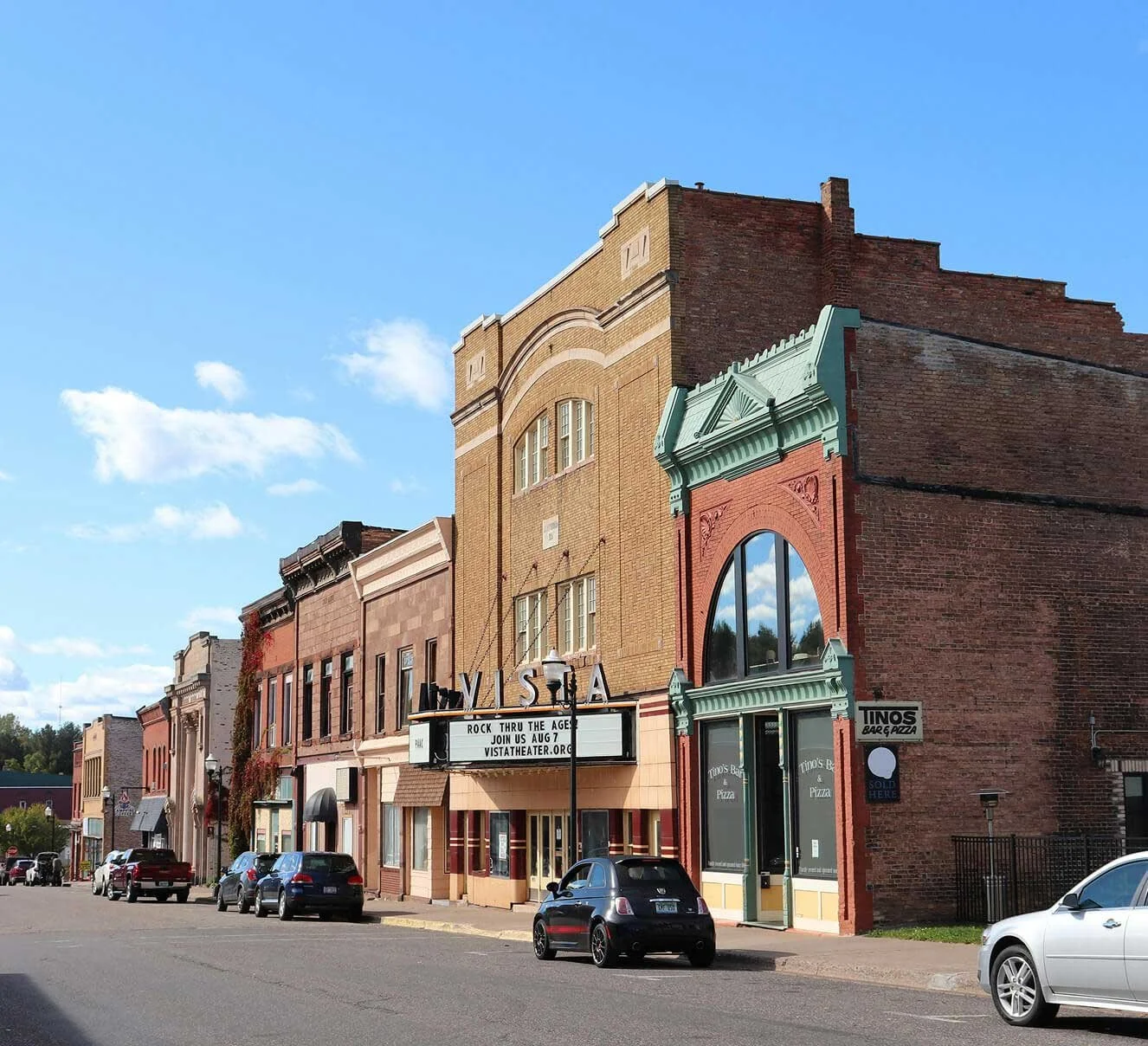 Street view of historic brick buildings with parked cars, a theater marquee, and a pizza restaurant named Tino's Bar & Pizza.