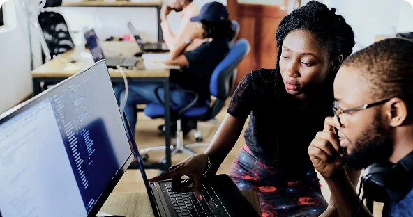 Two employees working together at a desk with a laptop and computer monitor in an office setting.