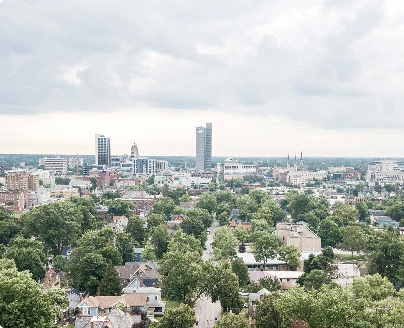 Aerial view of a cityscape with tall buildings, churches with steeples, and a neighborhood with many trees and houses under a cloudy sky.