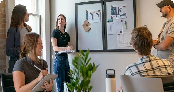Group of five young adults in a modern office, engaged in a presentation around a whiteboard with charts, notes, and a small 3D model, with one person speaking and others listening, taking notes, or observing.