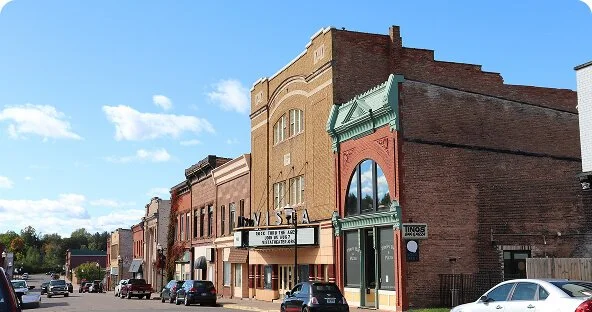 A street view featuring historic brick buildings with storefronts, parked cars, and a clear blue sky.