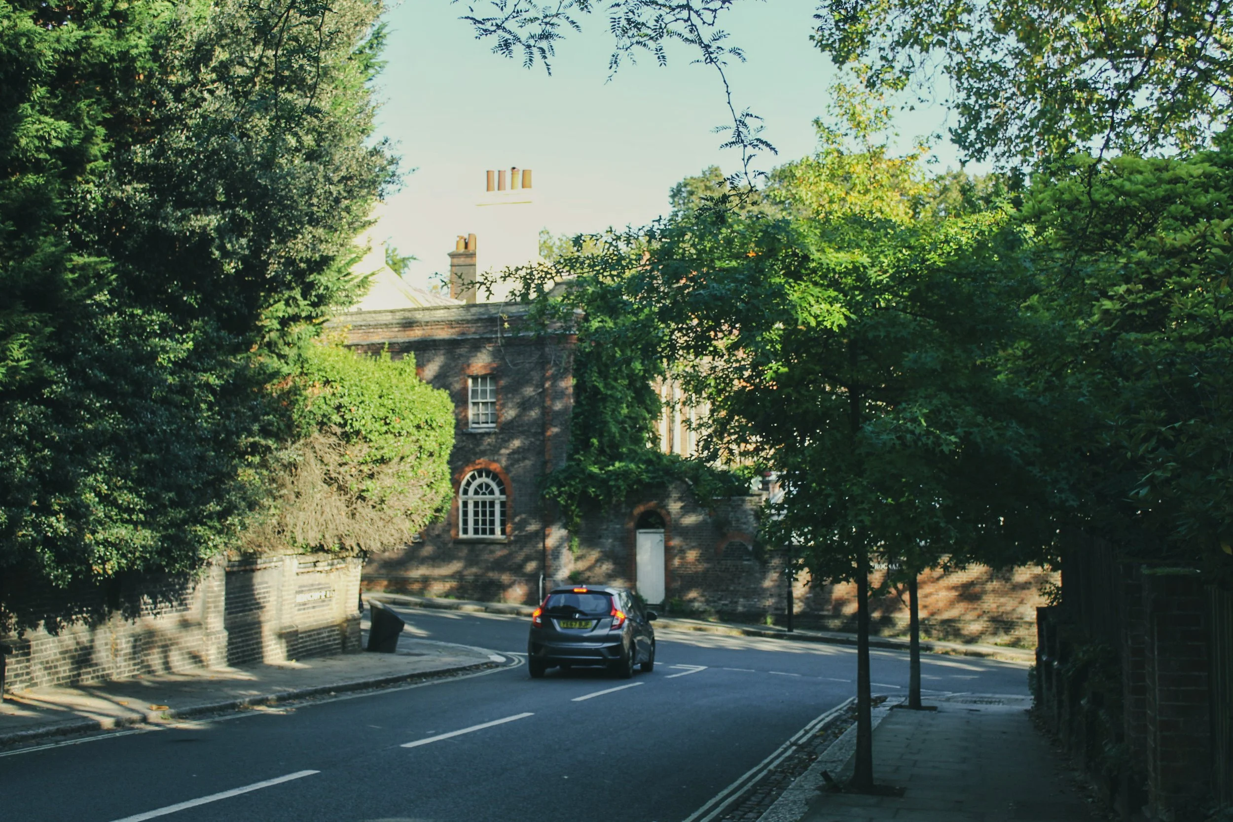 A quiet residential street with a car driving along a curved road, surrounded by lush green trees and brick houses with arched windows.