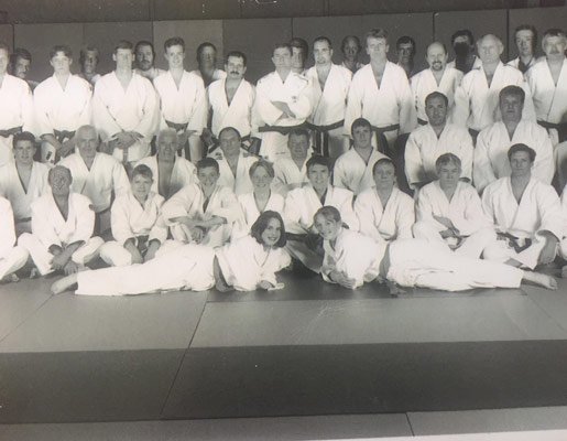 Group photo of people practicing martial arts in a dojo, dressed in white gis, with some seated and others standing, black and white image.