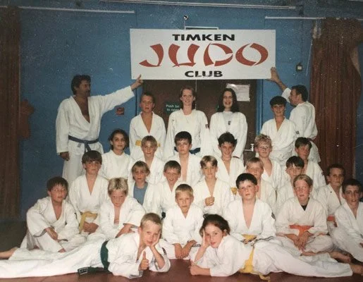 Group of children and adults in martial arts uniforms posing in a dojo in front of a sign that says 'Timken Ju Jitsu Club'.