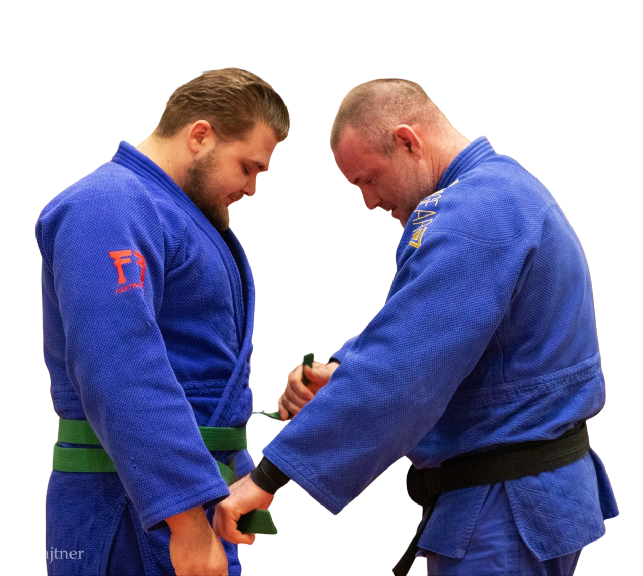 Two men in blue jiu-jitsu uniforms, one with a green belt and the other with a black belt, are standing face to face, locking hands and bowing heads in a gesture of respect.