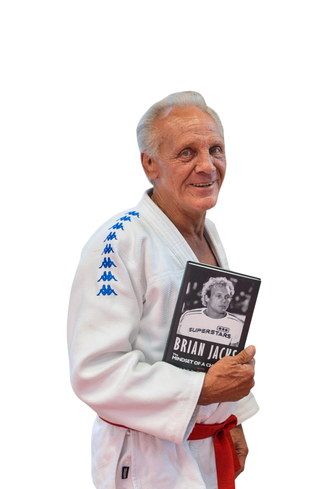 An elderly man wearing a white martial arts gi with blue logo on the shoulder, red belt, holding a book titled 'Brian Jacks: The Mindset of a Champion', smiling at the camera.