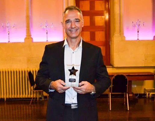 A man in a suit holding an award trophy in a decorated indoor setting with purple lighting and chairs in the background.
