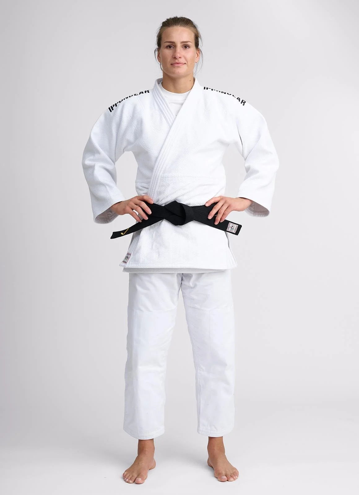 A woman standing in a white martial arts gi with a black belt, hands on hips, facing forward in a neutral pose.