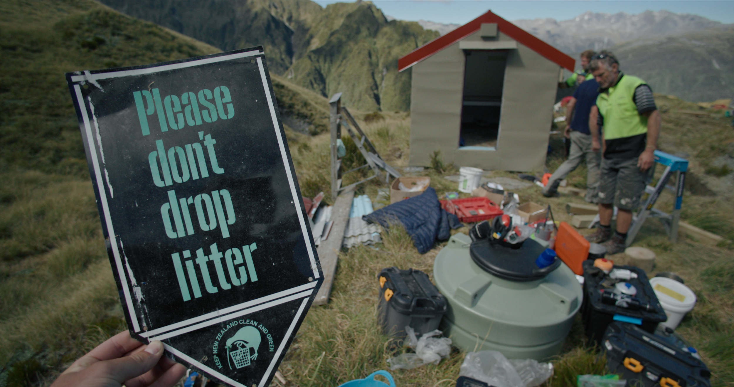 A person holding a sign that says 'Please don't drop litter' in a scenic outdoor area with mountains in the background. There are several people setting up or working at a site with various tools and equipment scattered around.