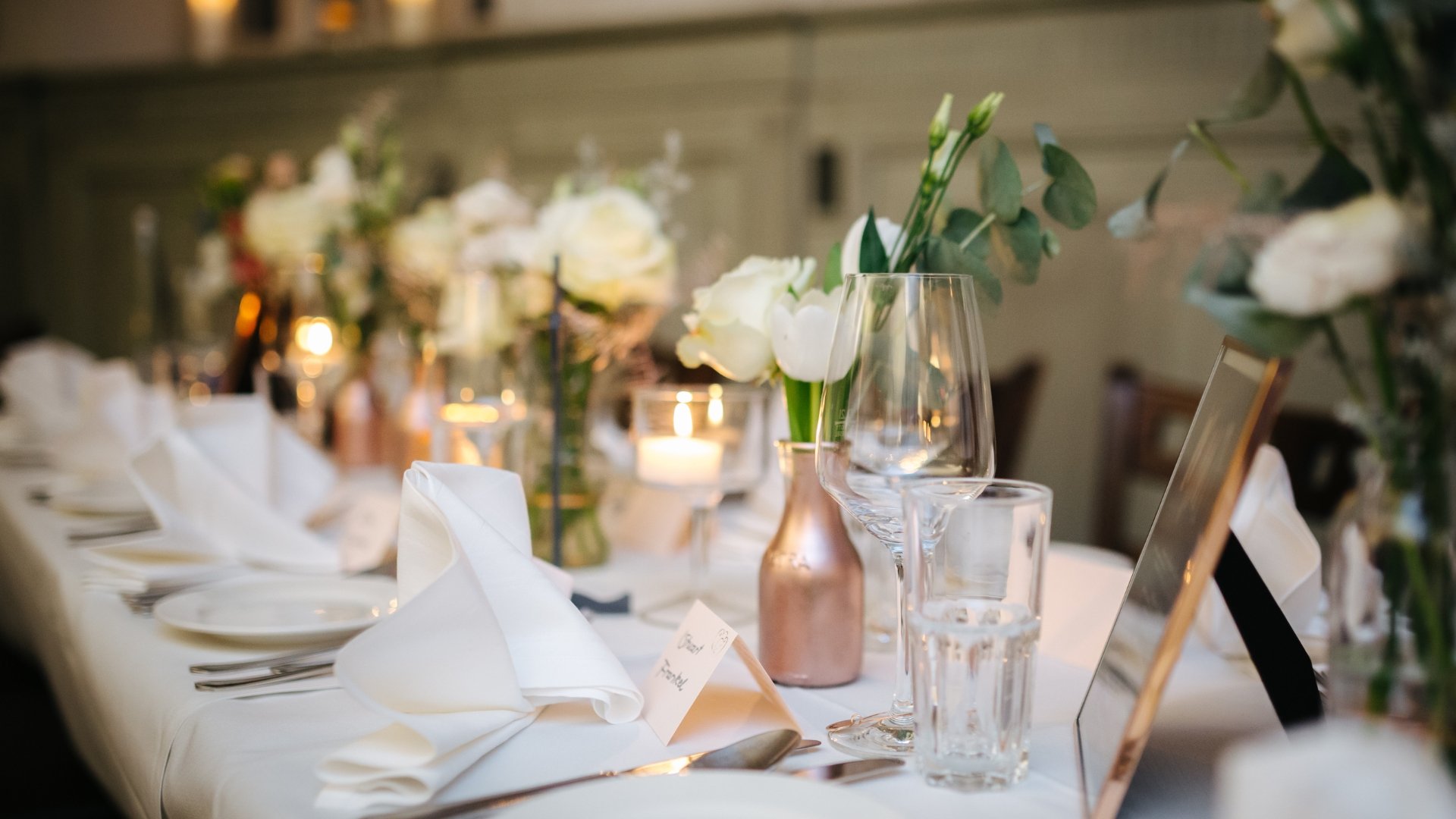 Elegant dining table set with white napkins, glassware, flower arrangements, and candles, ready for a formal event or celebration.