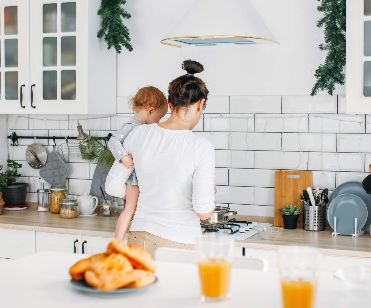 A woman and a child in a white kitchen, preparing food with Christmas decorations hanging from the cabinets and a plate of croissants and glasses of orange juice on the table in the foreground.