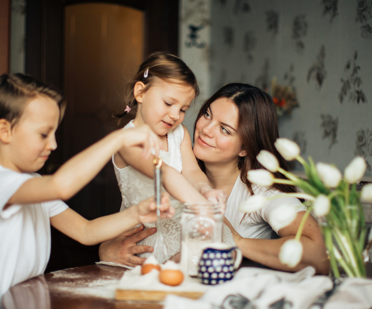 A woman helping two children with an activity at a table, flowers in the foreground, and a patterned wall in the background.