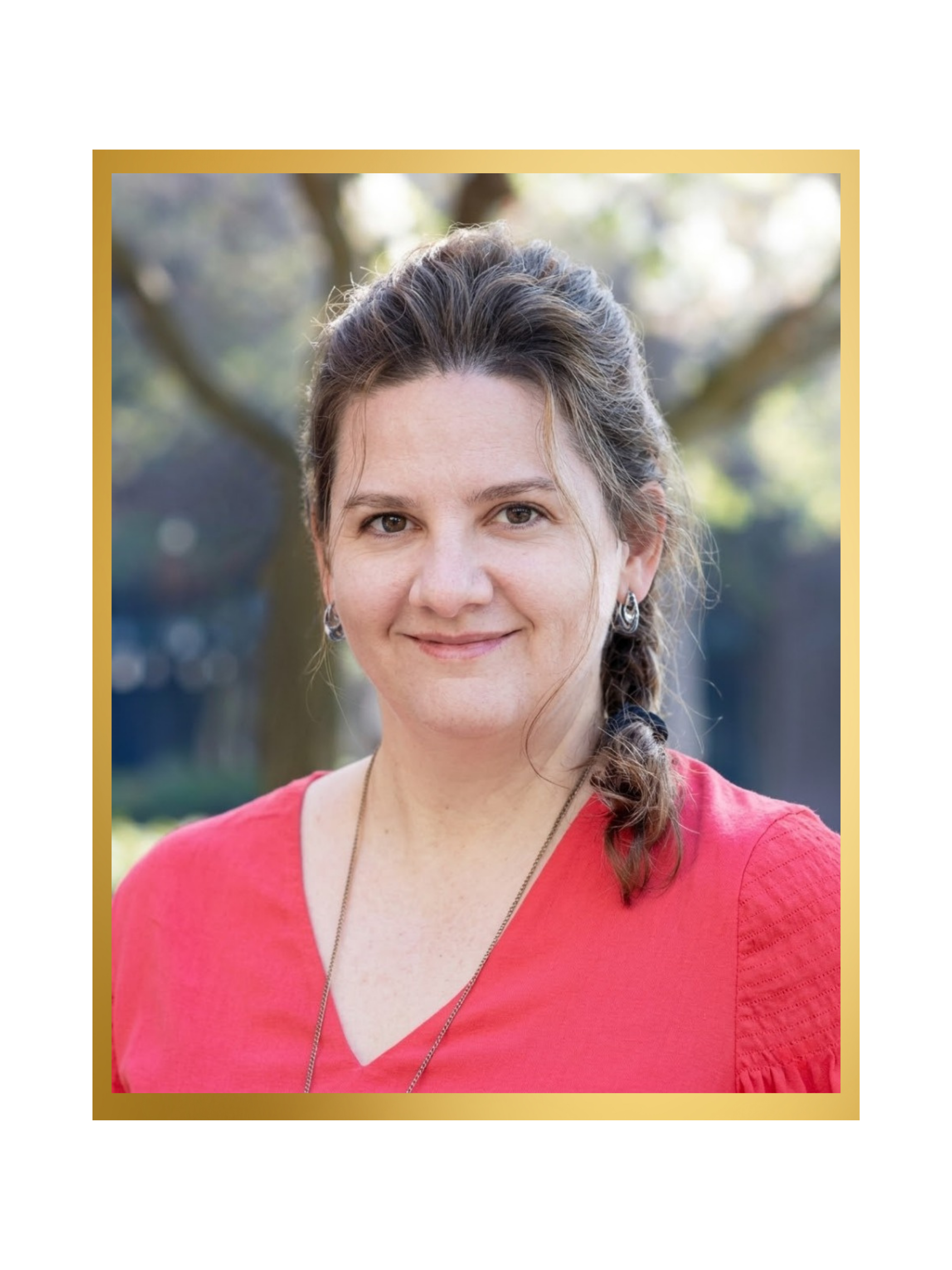 Portrait of a woman with brown hair, wearing a red top and silver earrings, outdoors with trees in the background.