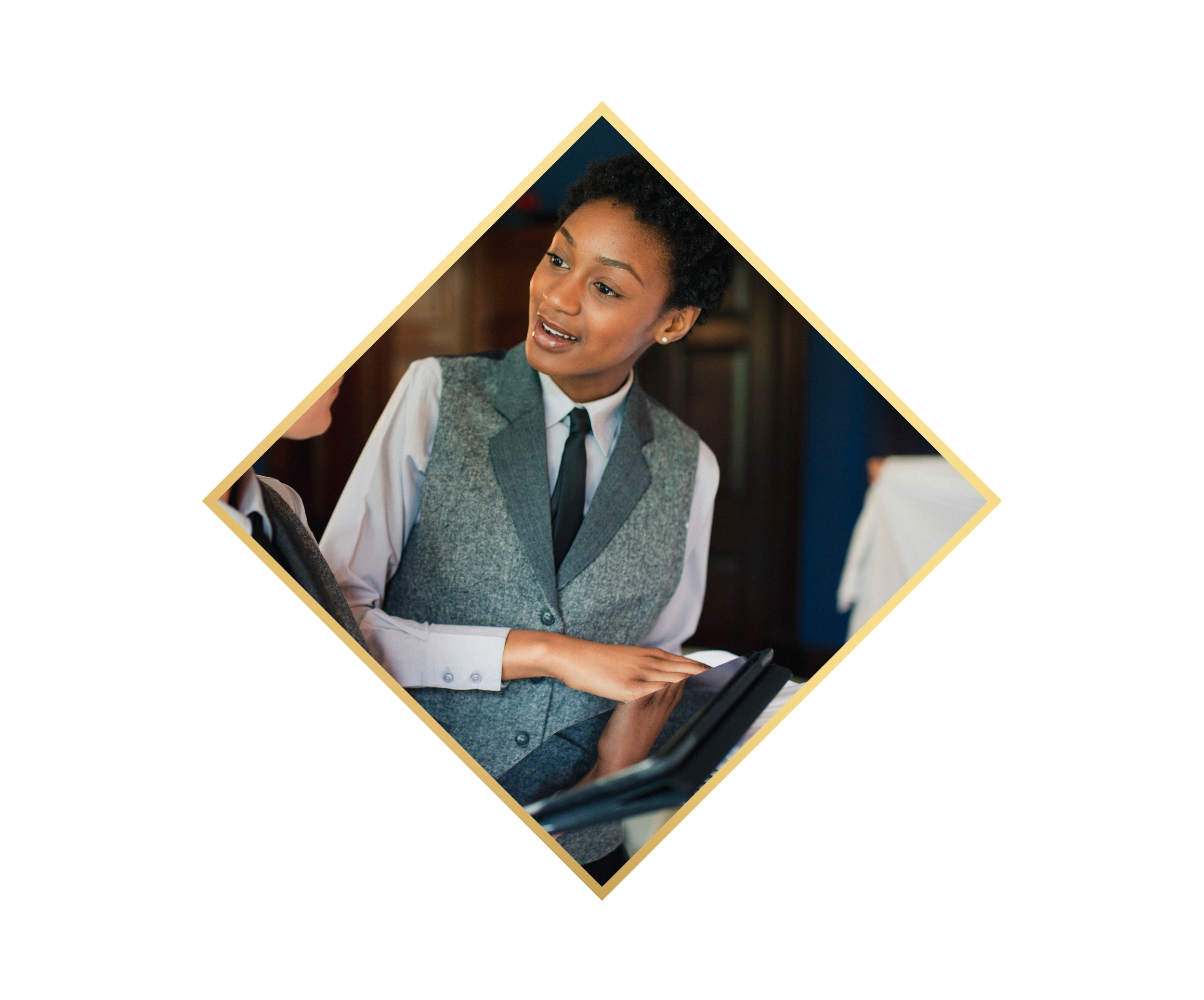 A woman in a grey vest and white shirt engaging in conversation with another person, holding a tablet.