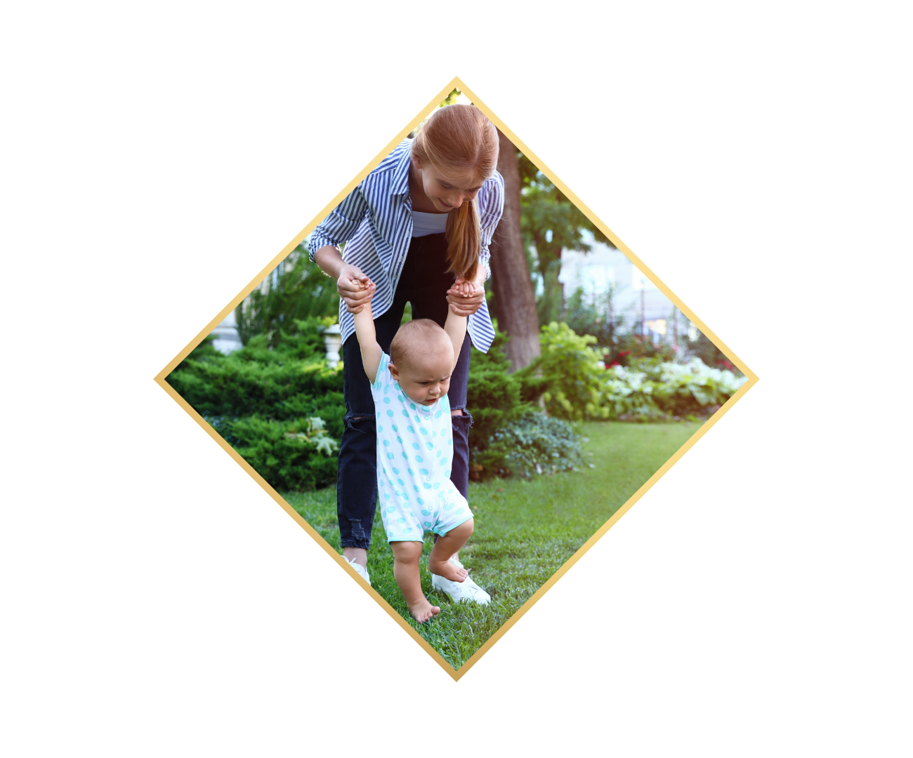 A woman is helping a young child learn to walk outdoors in a grassy park with trees and bushes, holding the child's hands for support.