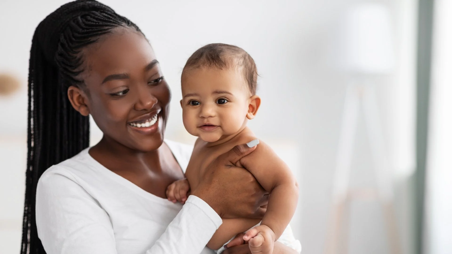 A woman with dark skin and long braided hair holding a smiling baby with light skin and short brown hair.