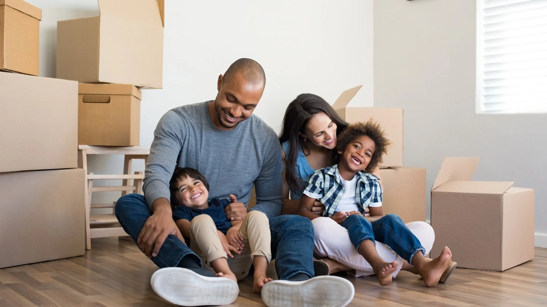 A family of four sitting on the floor of a room surrounded by packing boxes, smiling and enjoying each other's company.