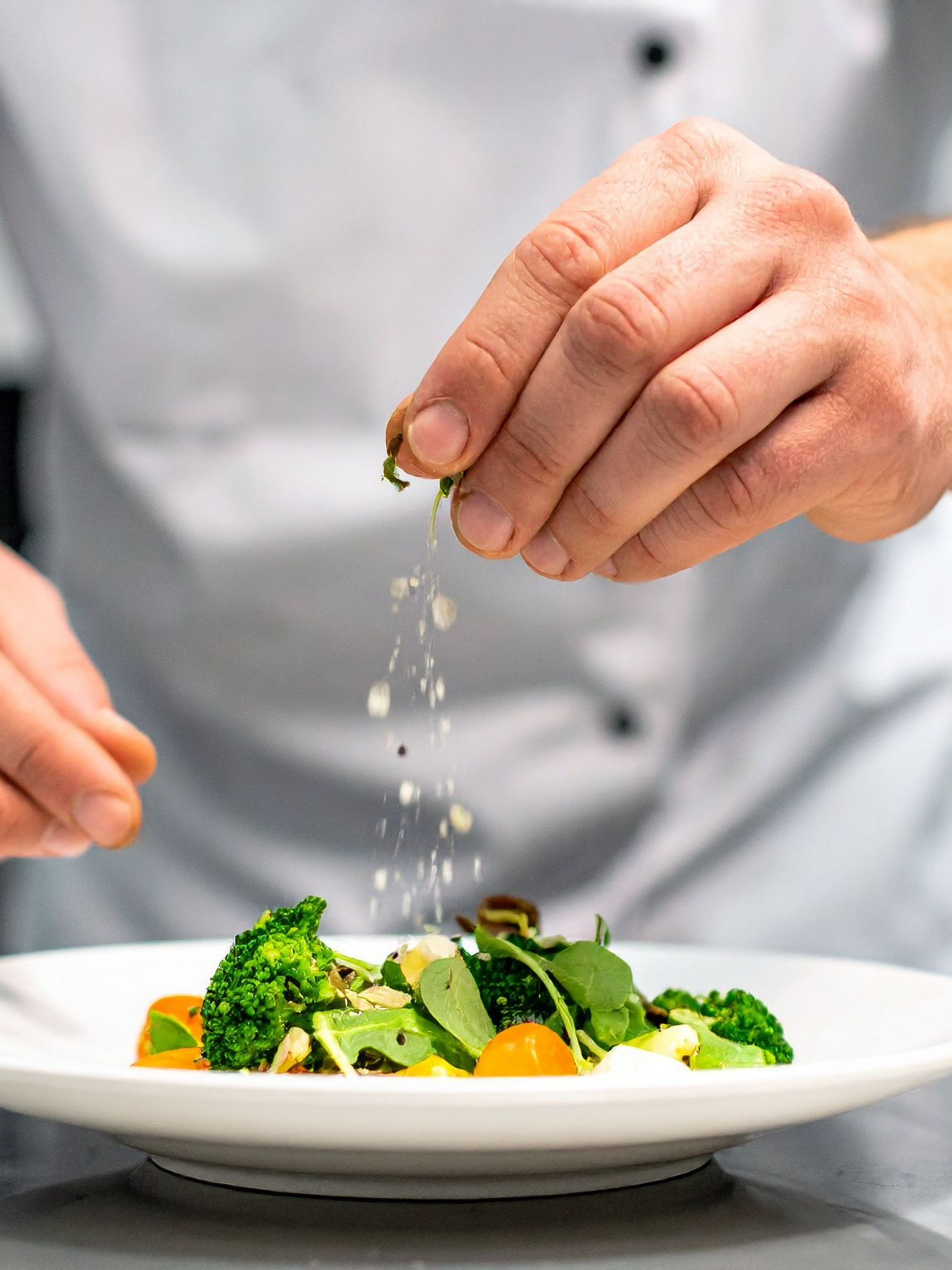 Person sprinkling seasoning or garnish onto a plate of mixed greens and vegetables.