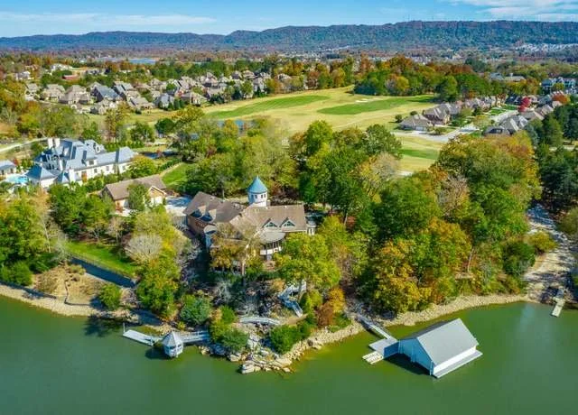 A scenic aerial view of a lakeside neighborhood surrounded by trees with colorful autumn foliage, a large house with a blue turret, a dock, and a boathouse, with a distant view of a golf course and residential area in the background under a clear sky.