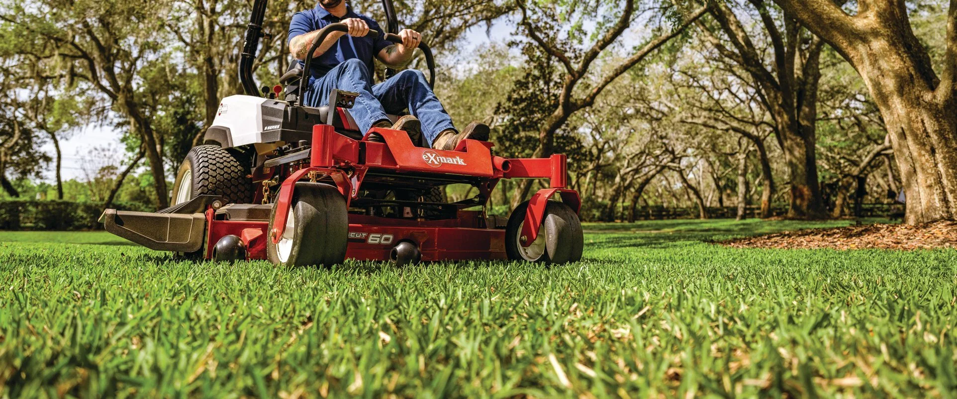 A person operating a red Exmark riding lawn mower on a well-maintained grassy yard with trees in the background.