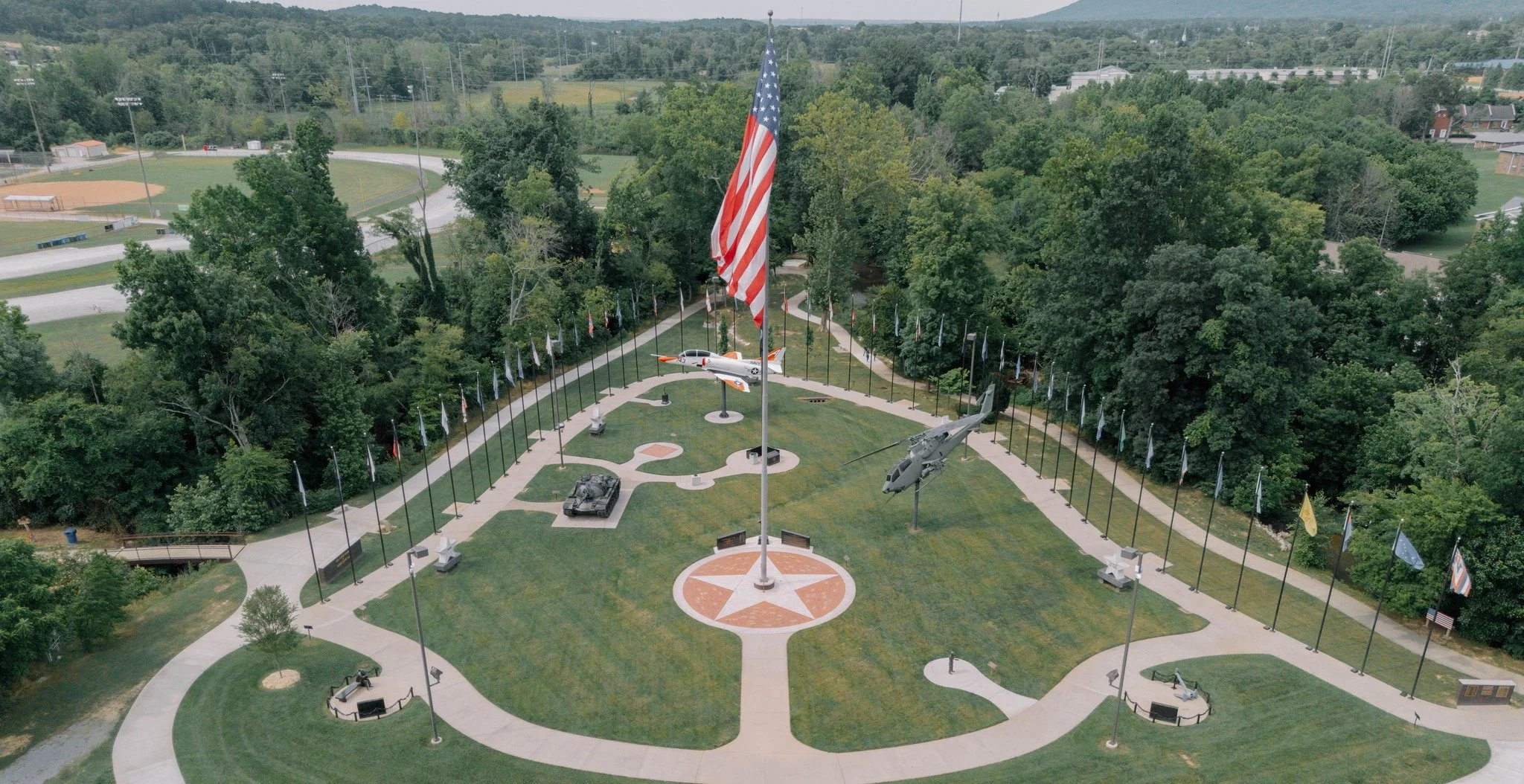 An aerial view of a military memorial park with a large American flag in the center, surrounded by flags, military vehicles, and planes, set within a park with trees and pathways.