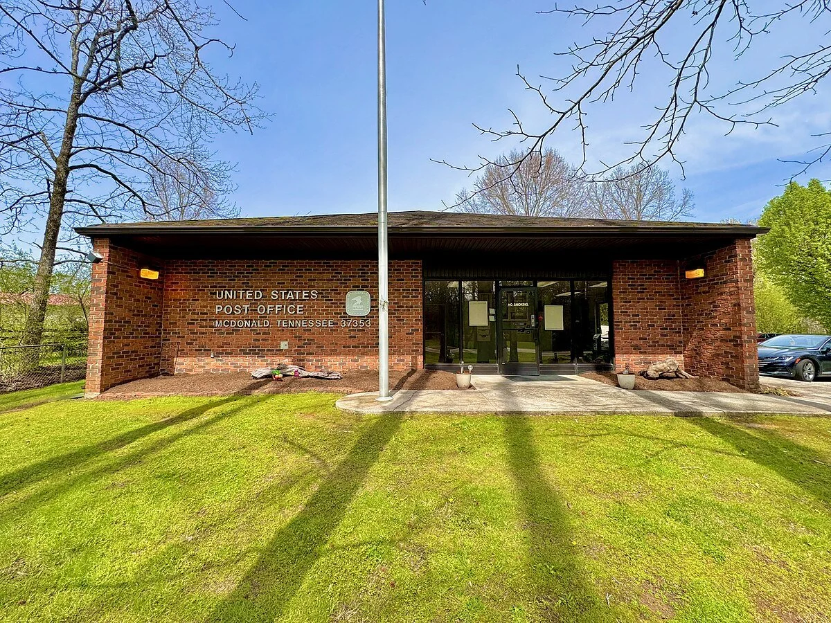 Front view of the United States Post Office in McDonald, Tennessee. The building is made of red brick with glass doors, and has a grassy area and parking lot in front. No people are visible.