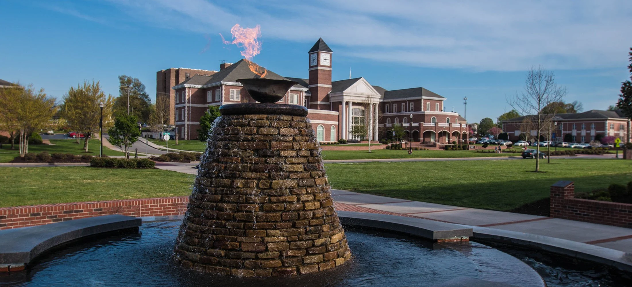 A brick fountain with a bowl on top, with water cascading down the sides, in a park area with grassy fields and trees. A large brick government or university building with a clock tower is visible in the background.
