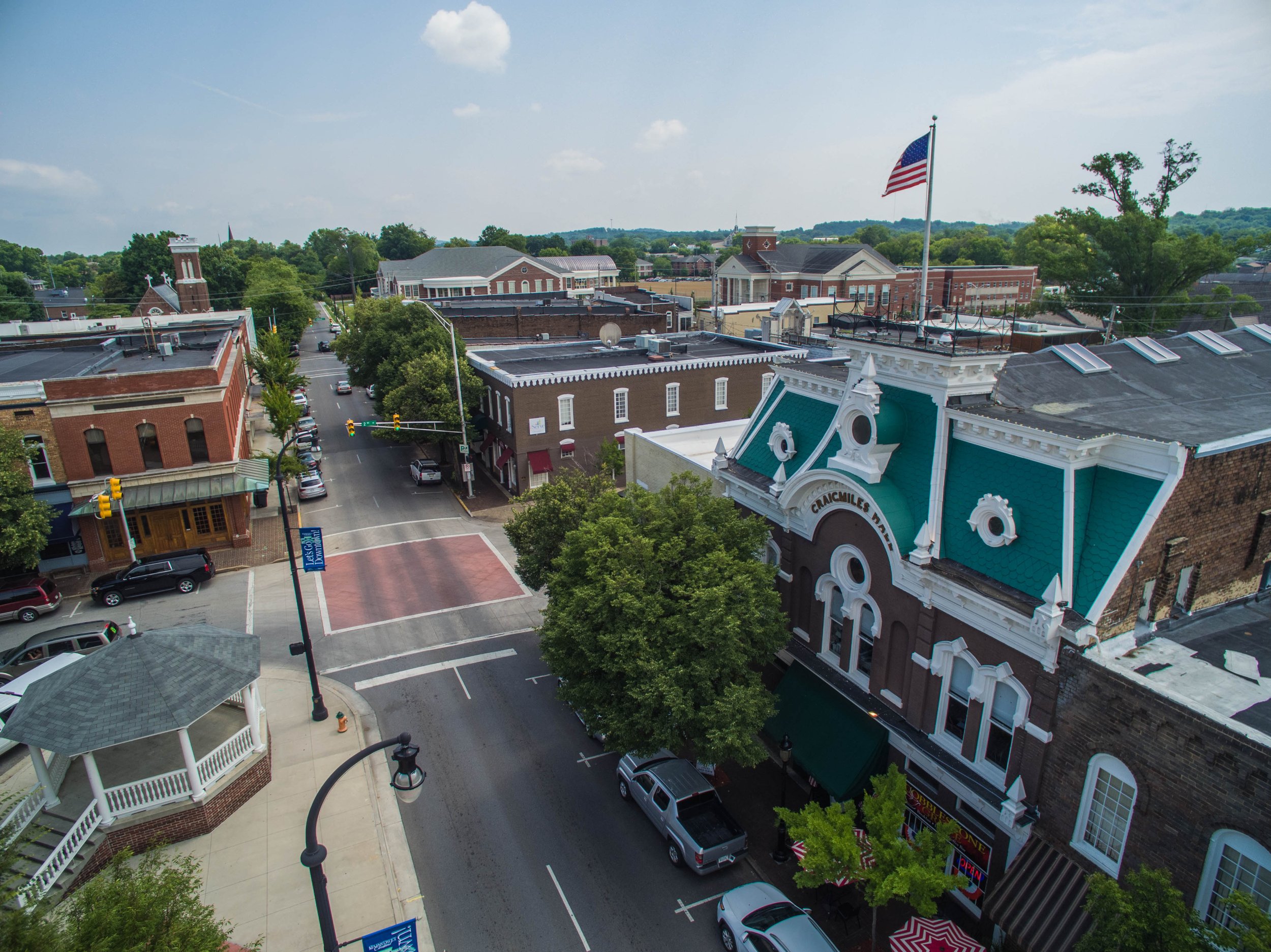 An aerial view of a small town street with brick buildings, cars parked along the road, and a large American flag flying on a flagpole on a building with a teal roof.