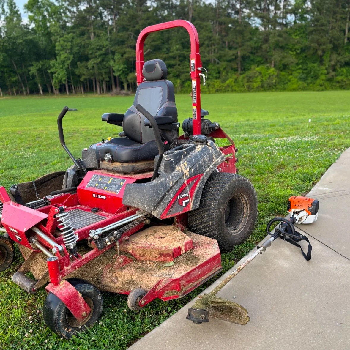 Red and black riding mower with grass and mud on the deck, parked on grass and concrete sidewalk near a wooded area, with an orange weed trimmer lying on the ground beside it.