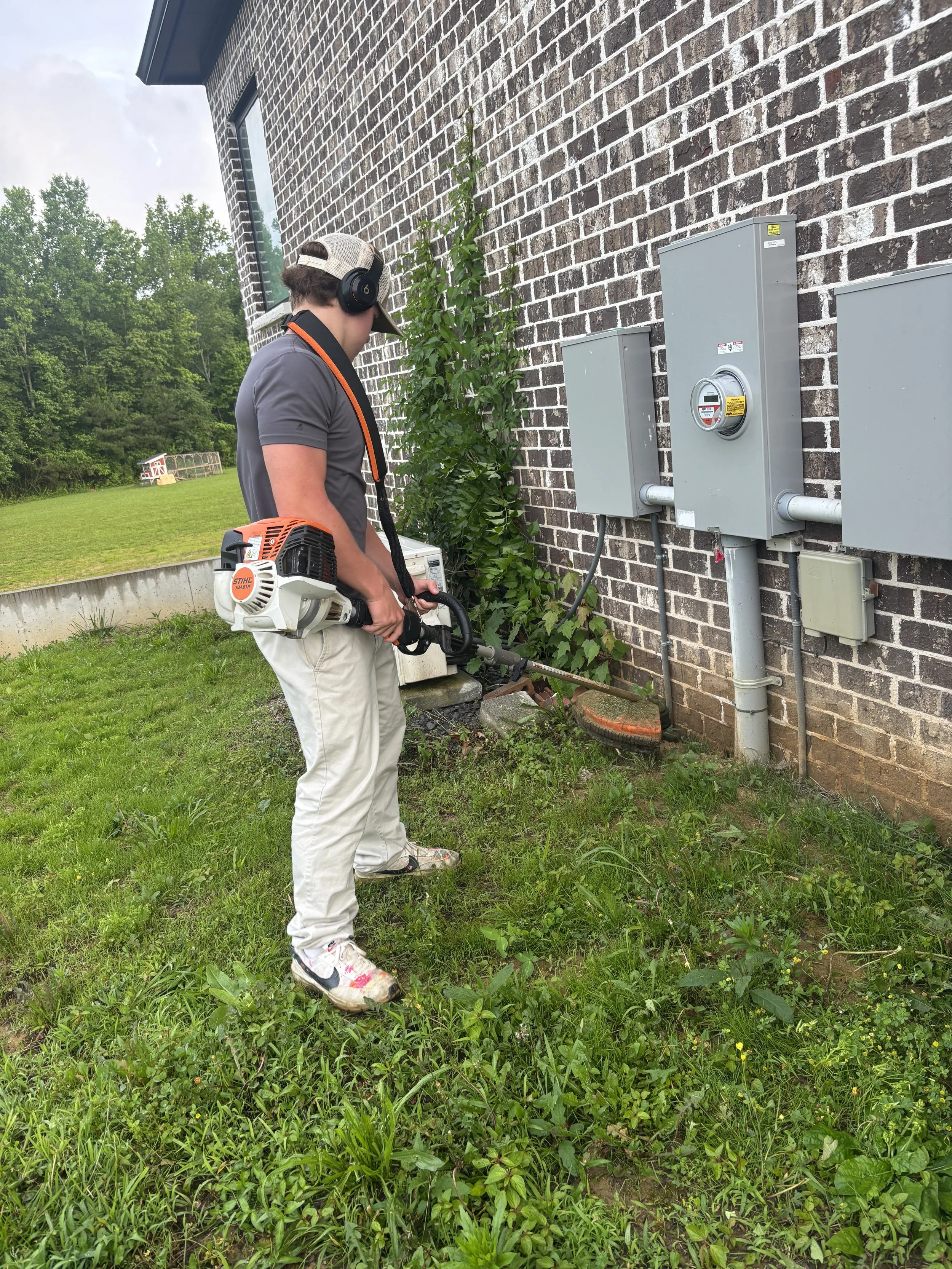 Person using a weed trimmer near the foundation of a brick building with electrical boxes on the wall, surrounded by grass and plants.