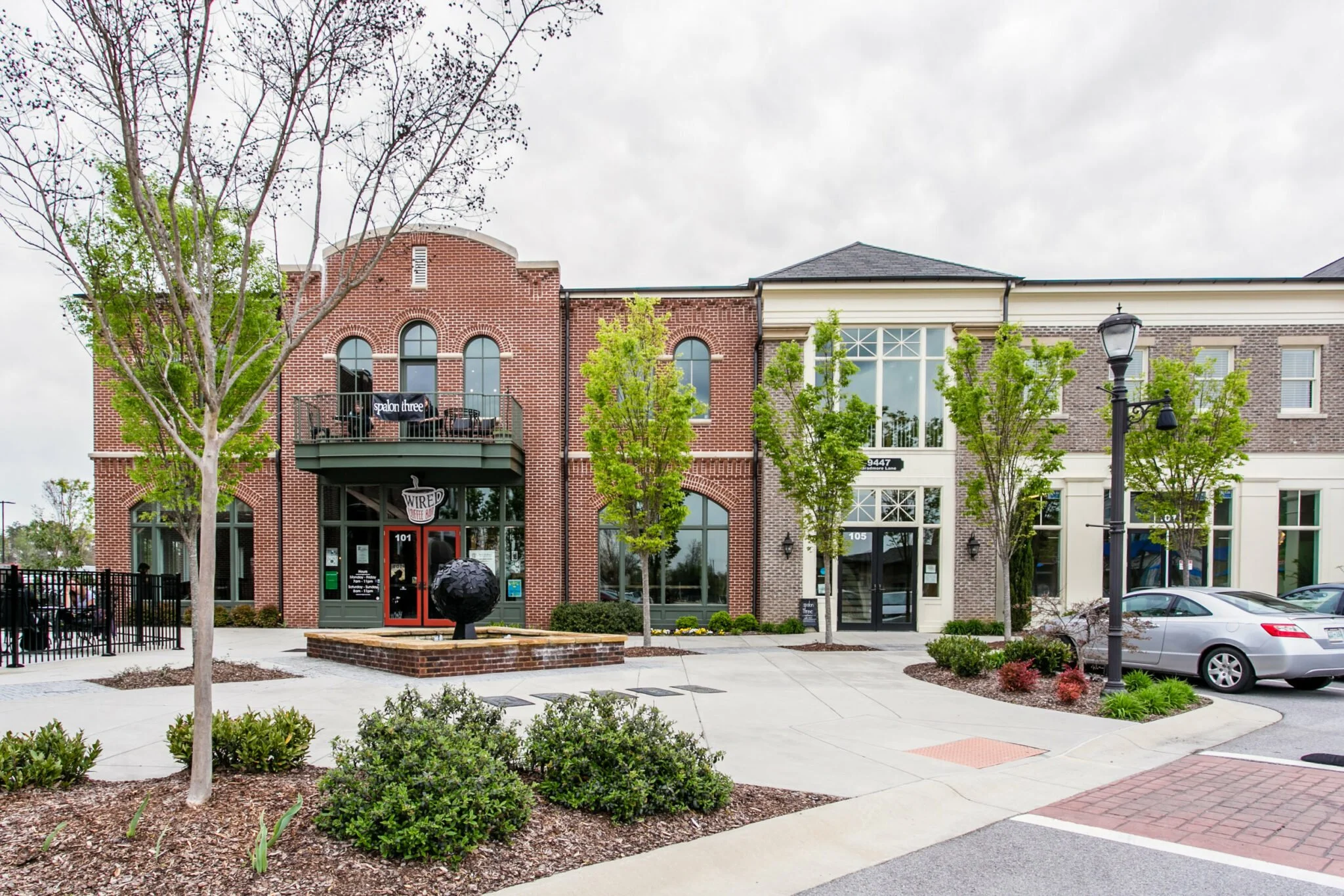 A modern commercial building with brick and white facade, featuring a cafe named Wired Coffee and a balcony with a sign for Spalon Three. The sidewalk in front has small trees, landscaped bushes, and a parking lot with a few cars.