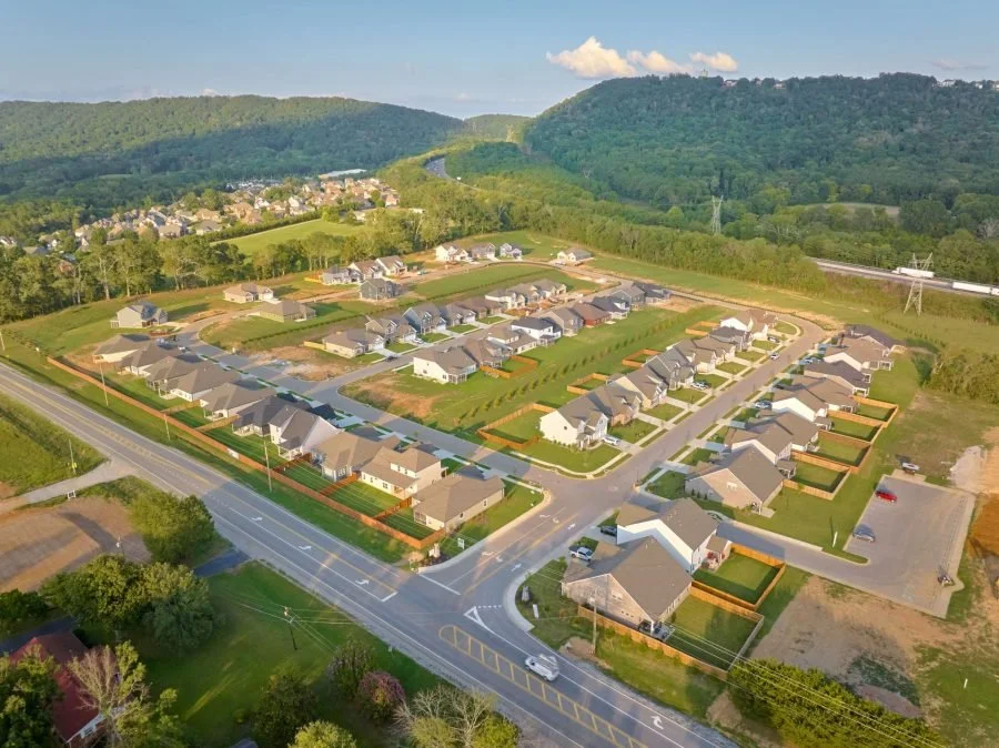 A bird's eye view of a suburban neighborhood with houses, streets, and green lawns surrounded by wooded hills and mountains.