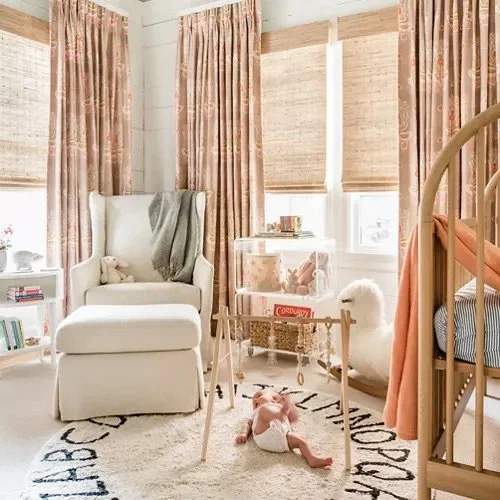 Cozy nursery with white armchair, pink floral curtains, window blinds, white plush llama, hanging toys, and a baby on a rug.