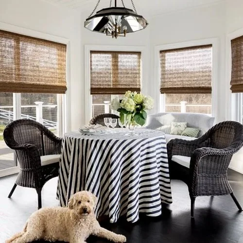 Sunlit dining area with a round table covered with a black and white striped tablecloth, surrounded by two wicker chairs and a white cushioned bench, with a floral centerpiece, a dog lying on the dark wooden floor, and three large windows with bamboo blinds.