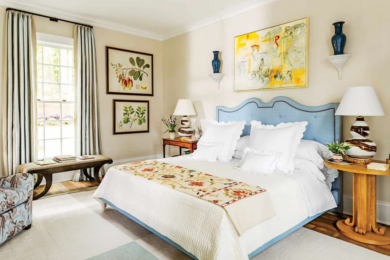 A cozy bedroom with a large bed featuring a light blue headboard and white pillows. There are two bedside tables with table lamps, floral decor, and artwork on the walls. A window with striped curtains allows natural light to brighten the room. A small bench and a floral sofa are also visible.