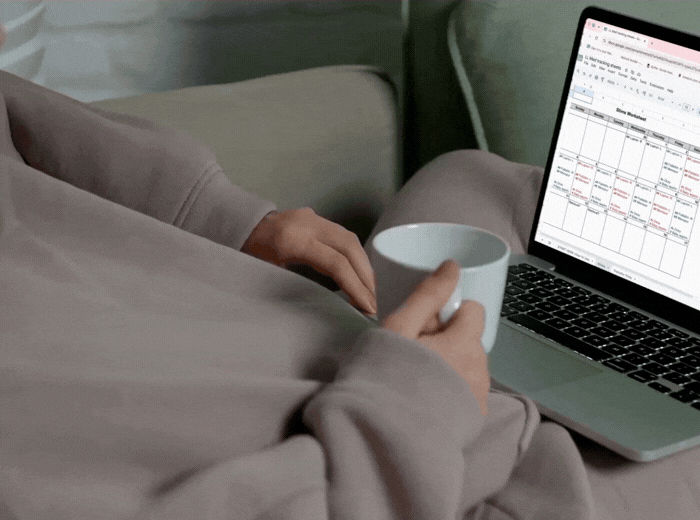 Person sitting on a couch holding a white coffee mug, with a laptop open showing a calendar or planning document.