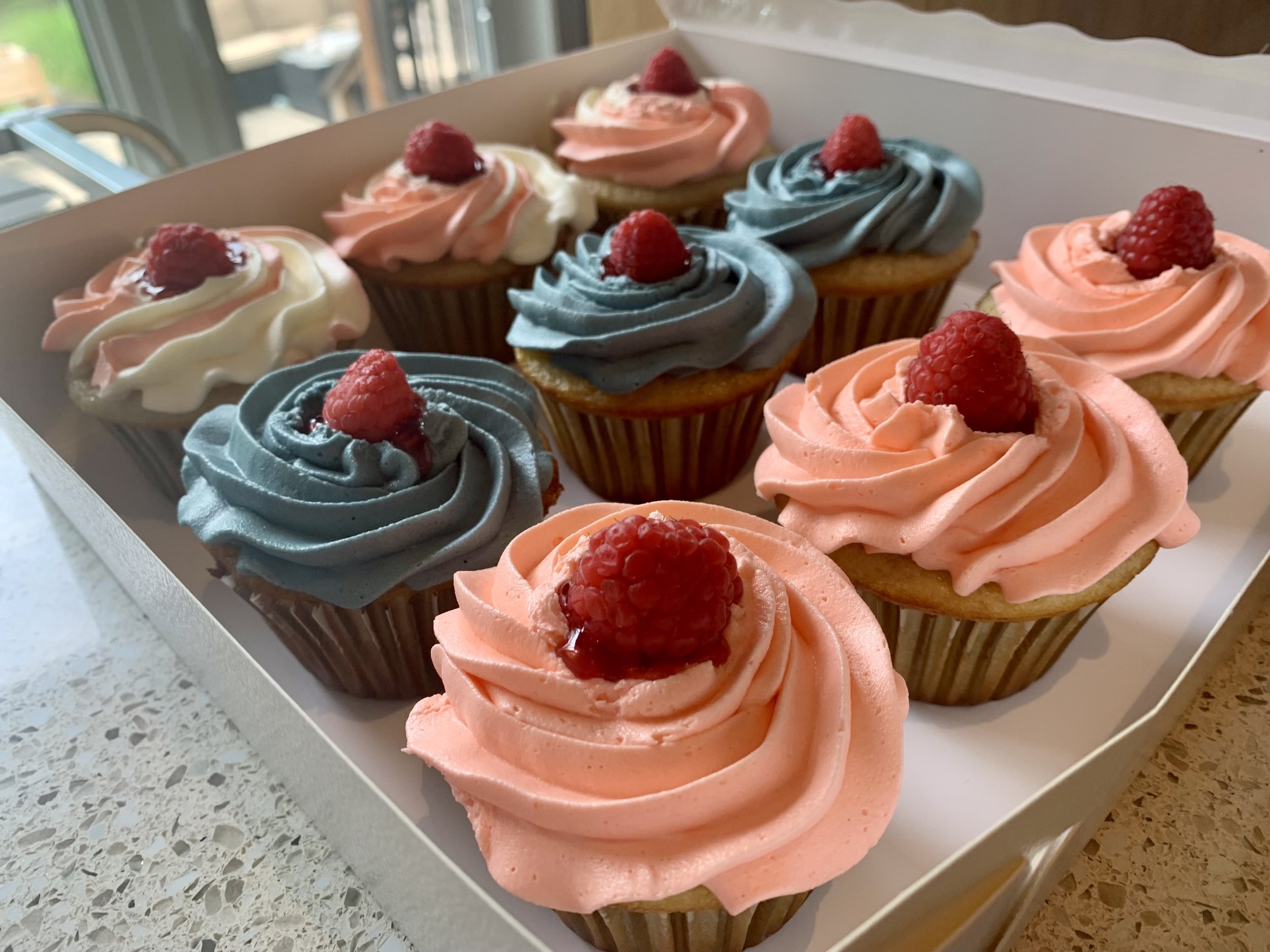 A box of twelve cupcakes with pastel-colored swirled frosting and fresh raspberries on top, arranged in pink and blue cupcake liners.