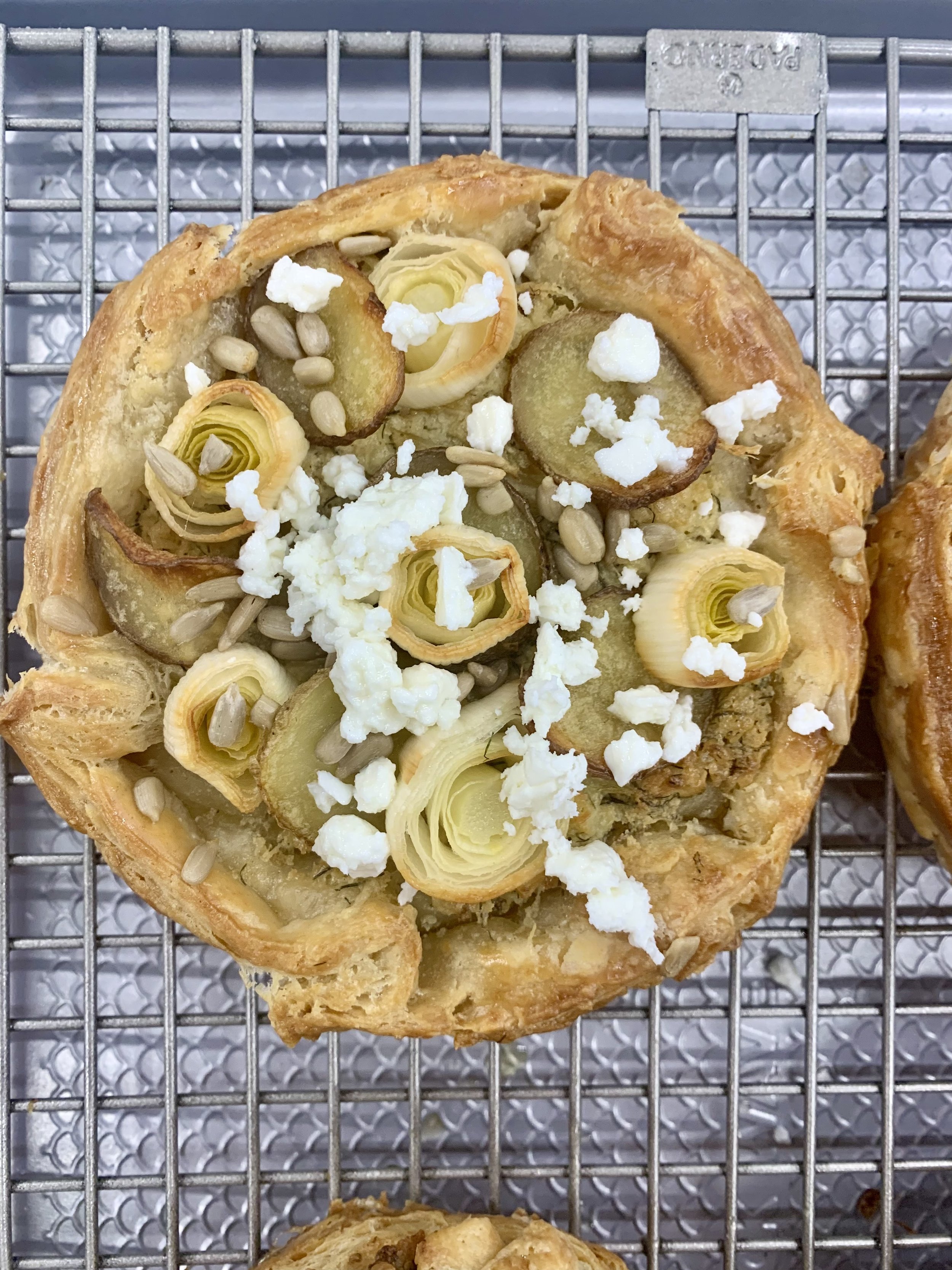 A savory galette topped with roasted leeks, sliced potatoes, sunflower seeds, and crumbled white cheese on a baking rack.