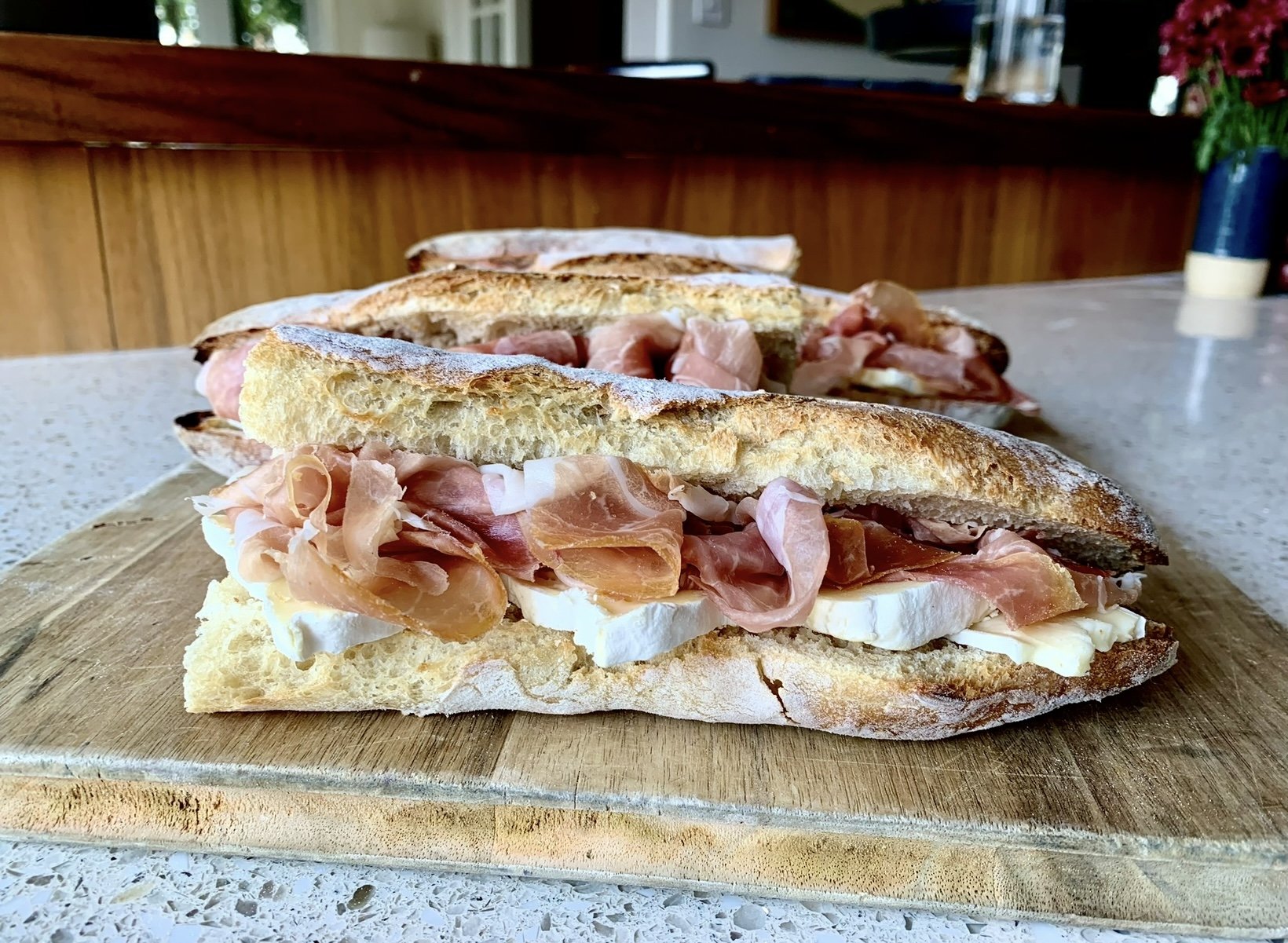 Close-up of prosciutto, mozzarella cheese, and baguette bread sandwich on a wooden cutting board, with additional sandwiches in the background on a kitchen counter.