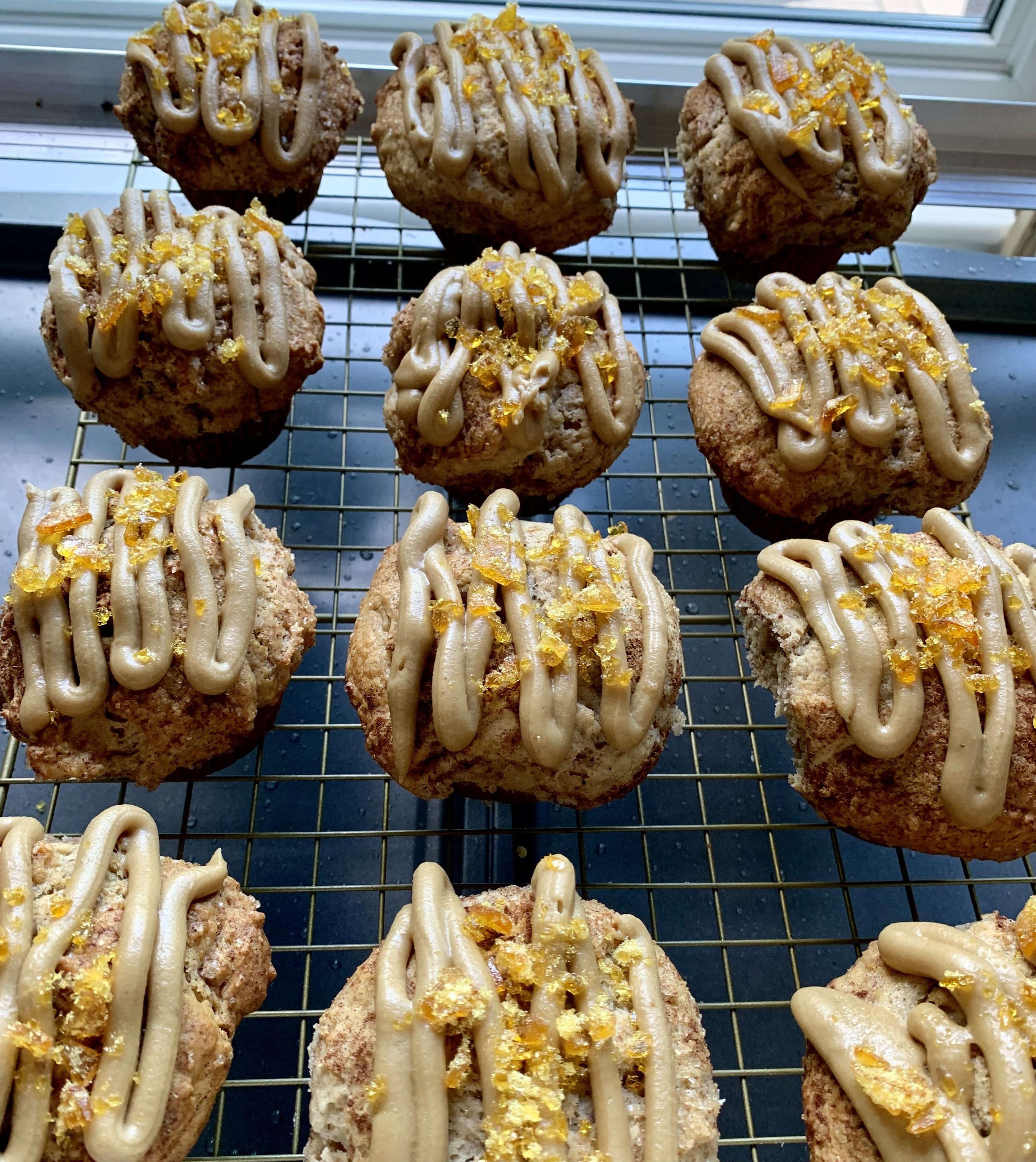 Baked muffins topped with vanilla frosting and yellow sprinkles on a cooling rack near a window.