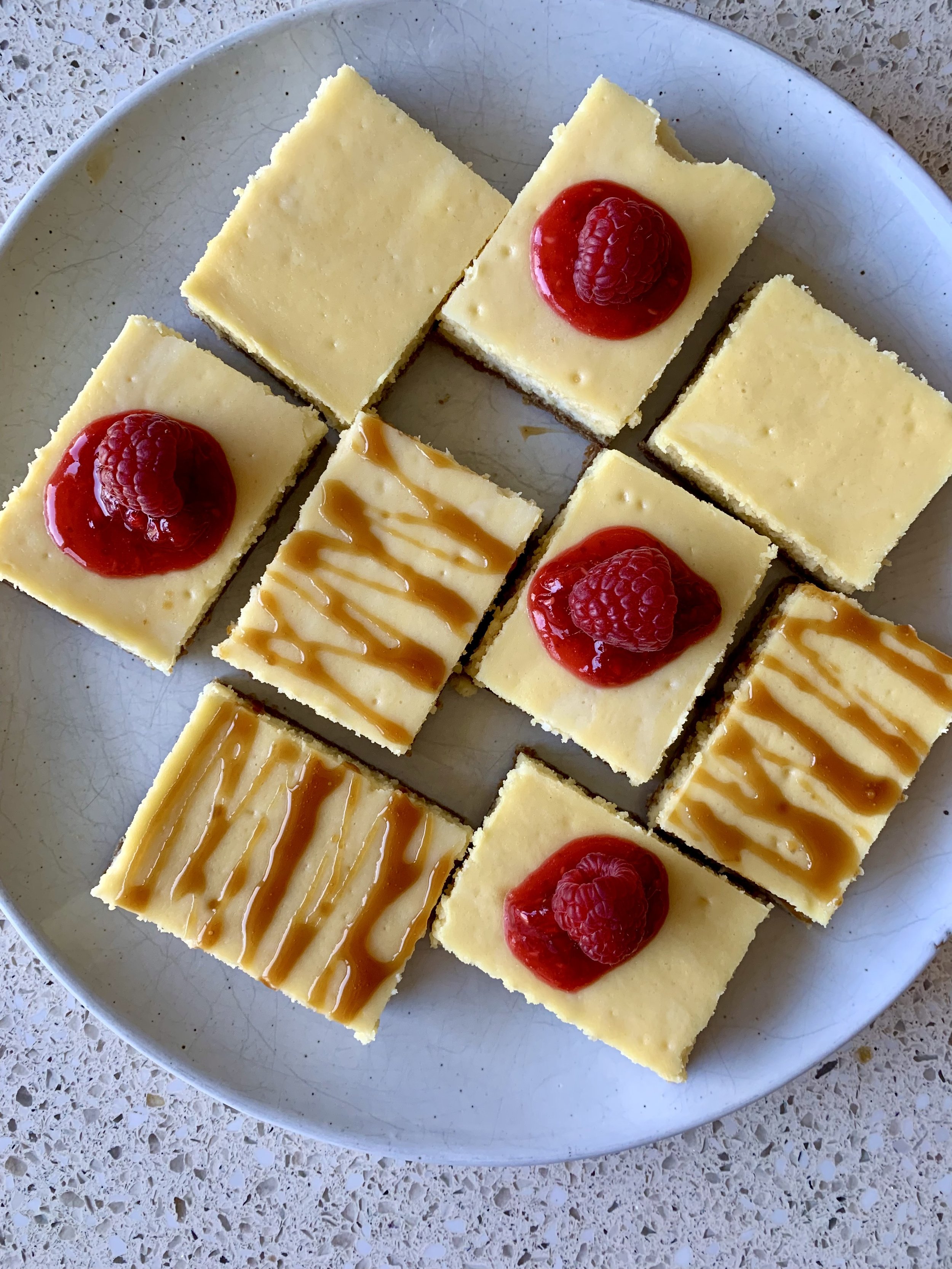 A white plate with nine small cheesecake squares decorated with raspberries and caramel or chocolate drizzle.