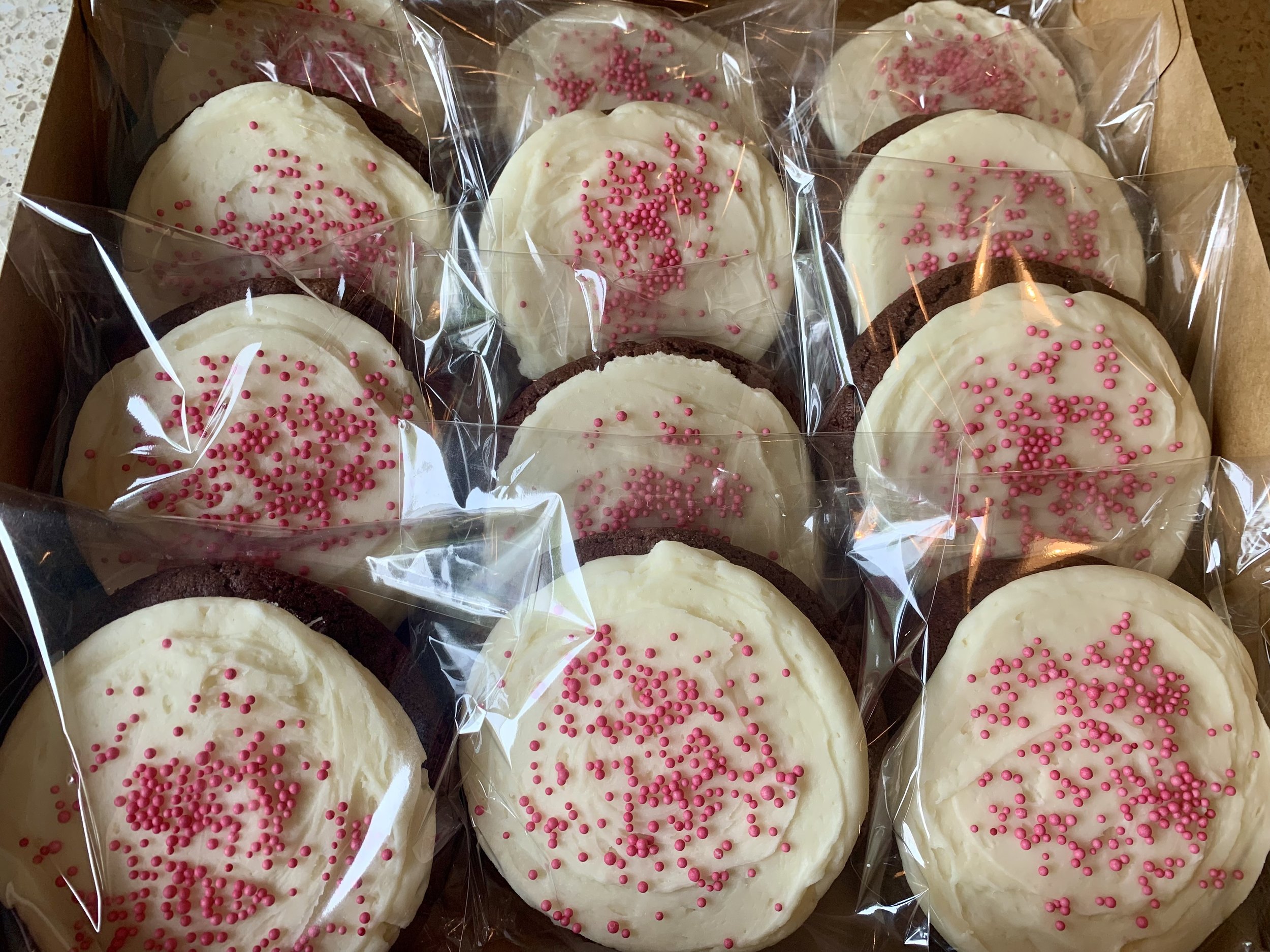 Box of decorated frosted red velvet cookies with white icing, pink sprinkles, and chocolate edges, individually wrapped in clear plastic.